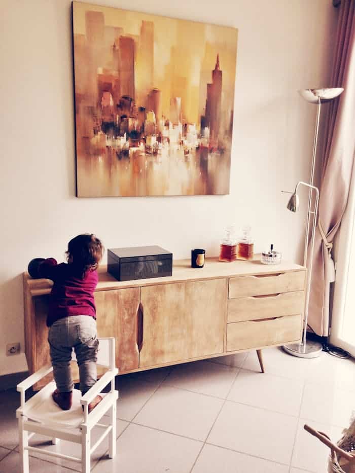 our-journey-03 Child standing on chair reaching a wooden cabinet in a modern living room with abstract painting.